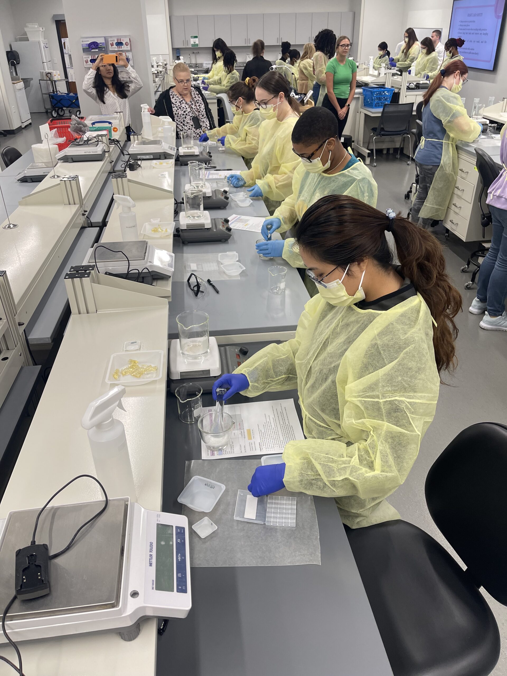 Photo showing a perspective focused on one row of students wearing yellow lab gowns and face masks are seated in a brightly lit laboratory classroom. Laboratory equipment and materials are arranged on the long white tables with black swivel chairs.
