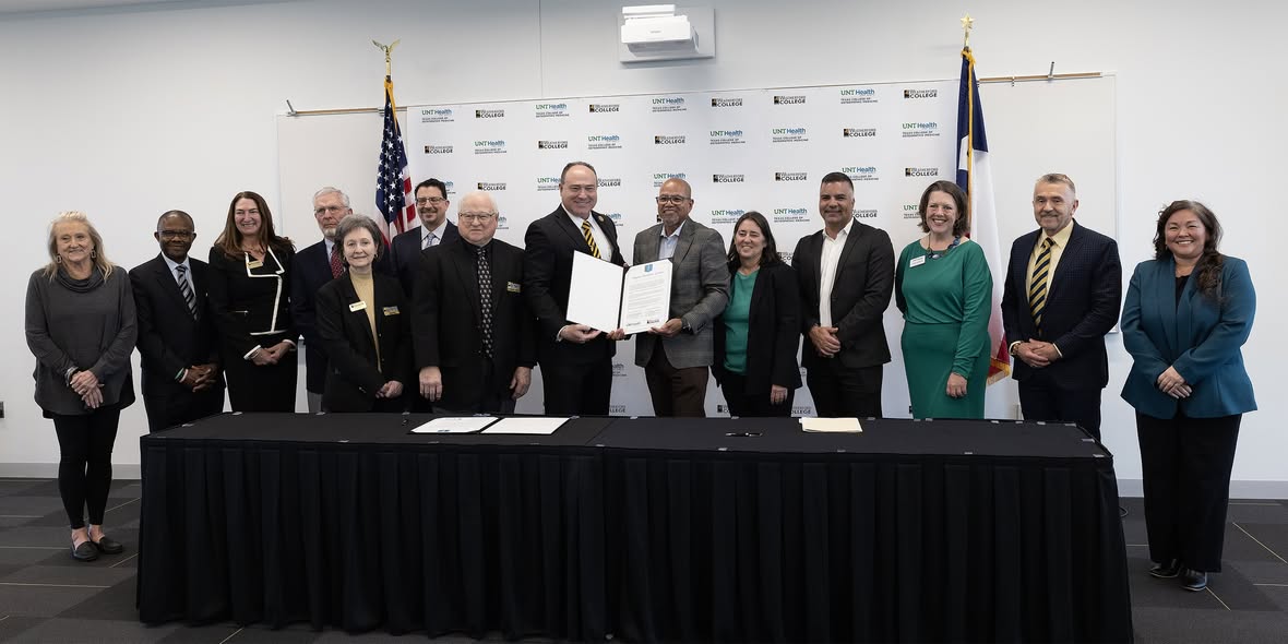 A large group standing behind their official signing ceremony table, representing both Weatherford College (on the left) and UNT Health Texas College of Osteopathic Medicine (on the right), celebrates their signed agreement. Full long horizontal photo of fourteen people standing behind their official signing ceremony table, representing both Weatherford College (on the left) and UNT Health Texas College of Osteopathic Medicine (on the right), celebrates their signed agreement. In the middle, both presidents, Dr. Tod Allen Farmer from Weatherford College and Dr. Kirk Calhoun from UNT Health, are holding up the document in an open black folder together. Behind them, on a wall, is a banner displaying both schools' repeated logos in a stair-stepped fashion in the background.