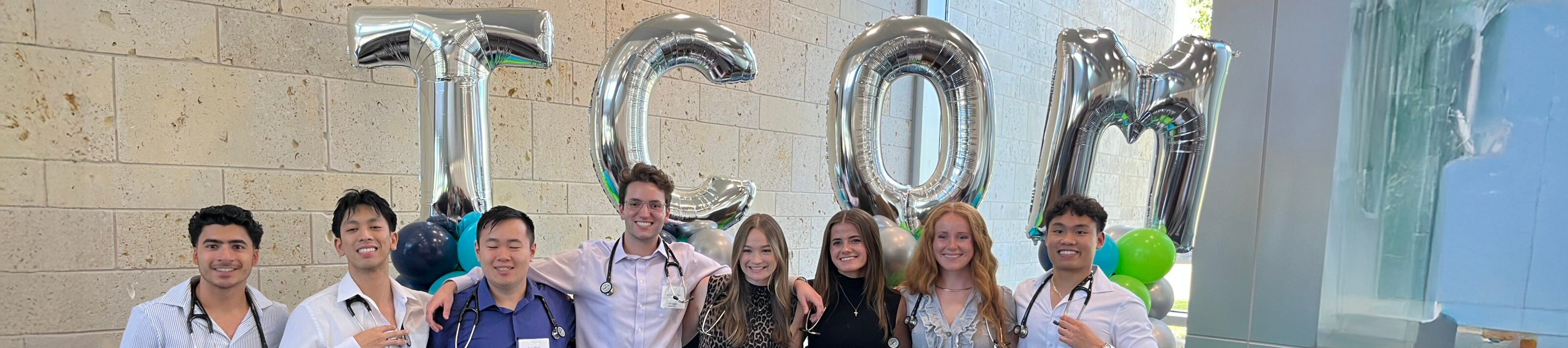 Top cropped photo banner of a group of 8 UNT Health smiling medical students wearing stethoscopes pose together in front of large balloon letters spelling “TCOM.” Top horizontal, narrow-cropped photo banner of a group of 8 UNT Health smiling medical students wearing stethoscopes pose together in front of large balloon letters spelling “TCOM.”