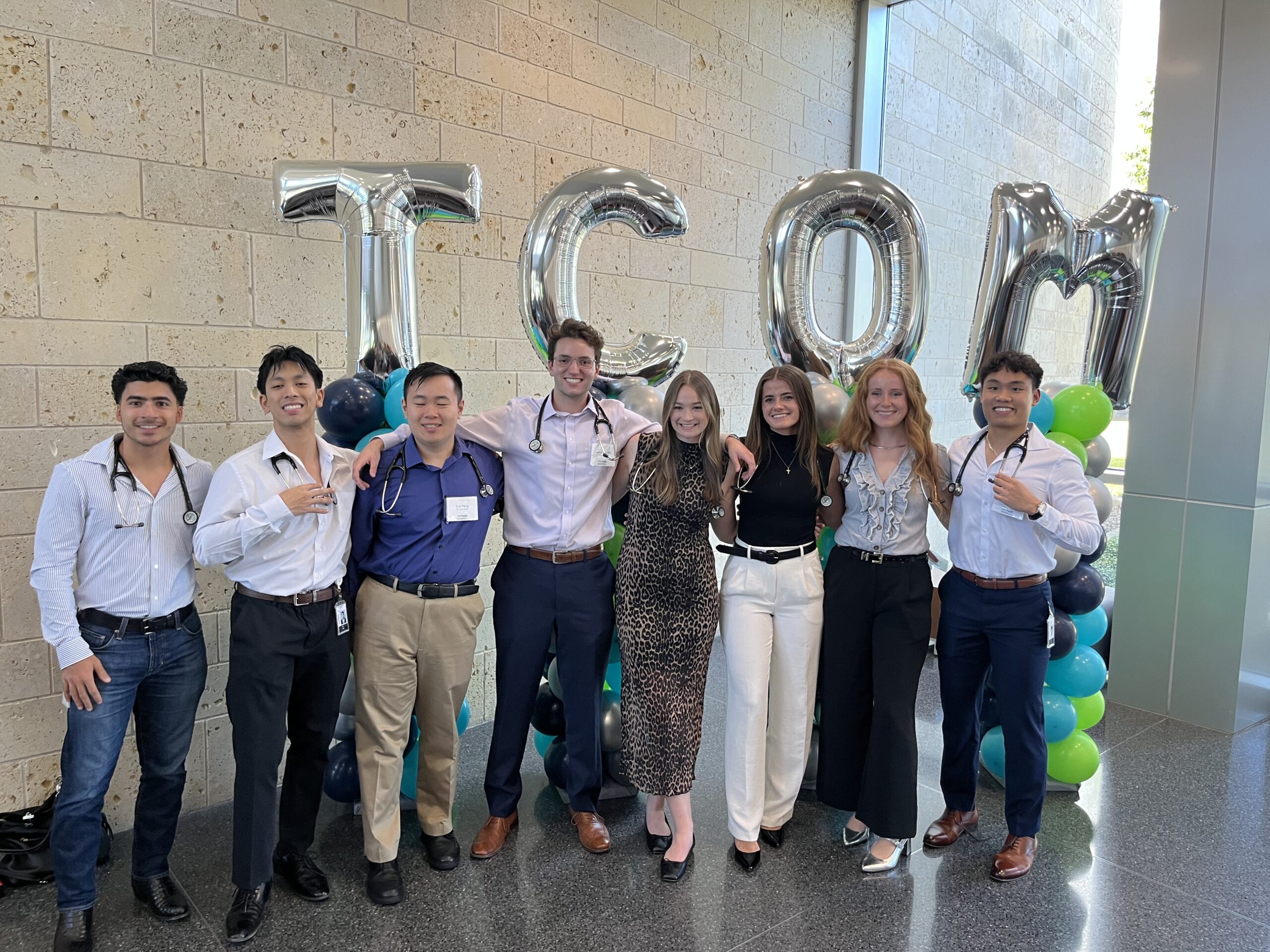  Full photo of a group of 8 UNT Health smiling medical students wearing stethoscopes pose together in front of large balloon letters spelling “TCOM.”