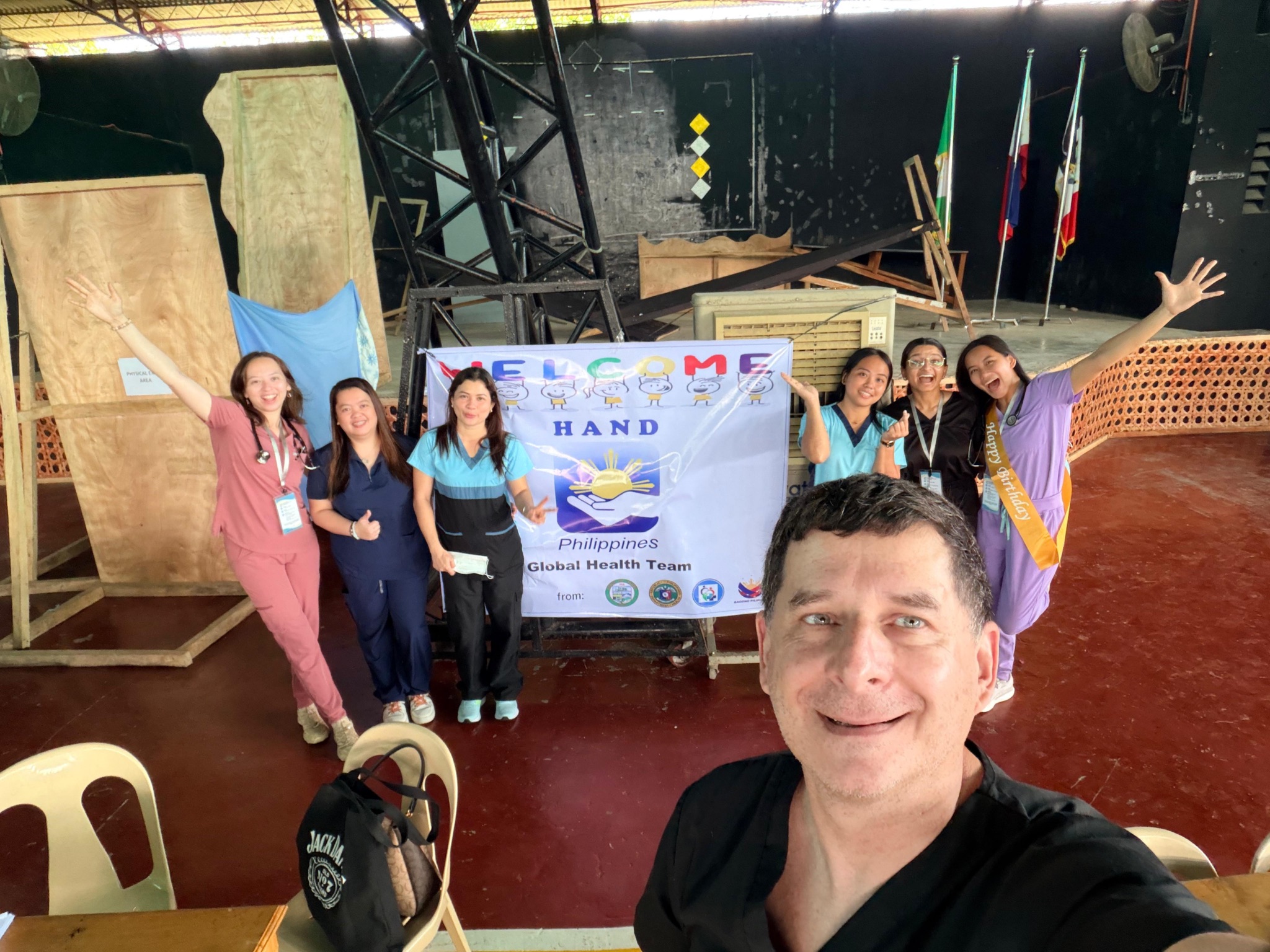Dr. Thomas Shima takes a selfie with UNT Health TCOM students and local Philippine medical staff celebrating in front of a HAND Philippines Global Health Team welcome banner during the Bohol medical outreach mission.