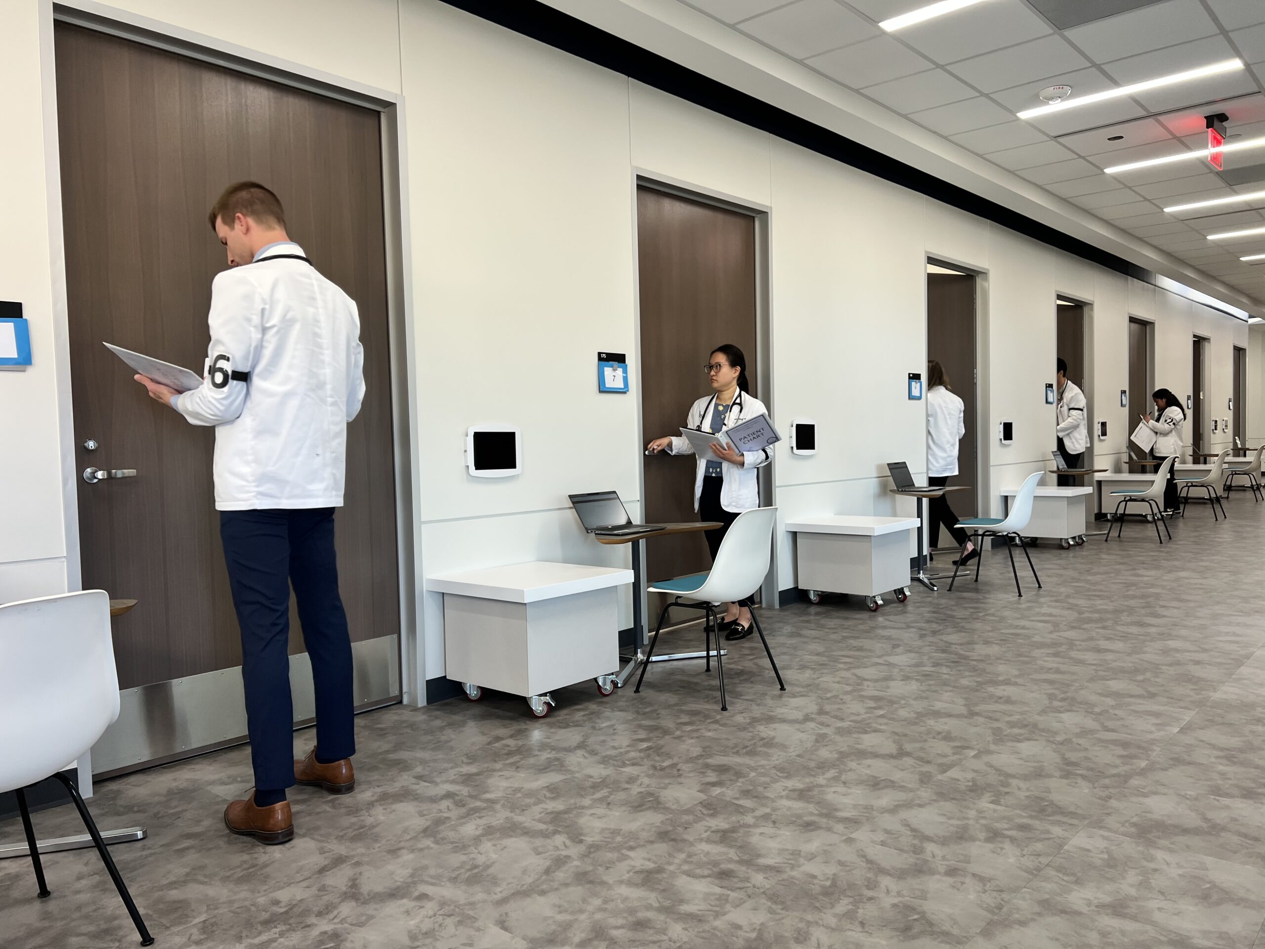 Photo of 5 UNTHSC TCOM medical students lining up in a hallway entering numbered rooms to interact with their training