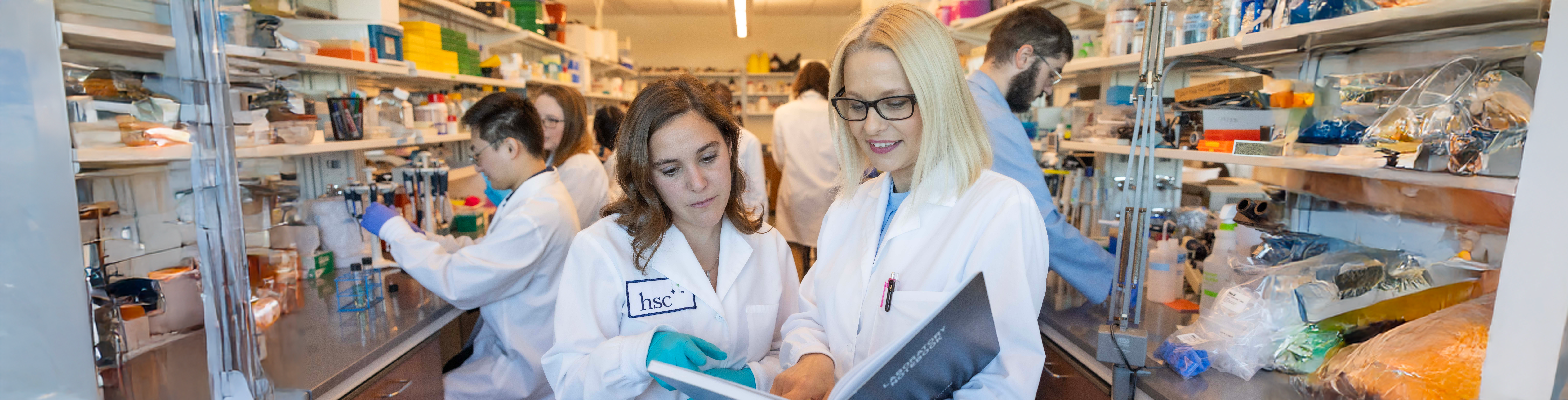 Top horizontal cropped photo of two female medical student researchers in white medical jackets looking at a bookbinder. Behind them are other student and faculty researchers handling test tubes and equipment sitting at opposing counters positioned on either side of them.