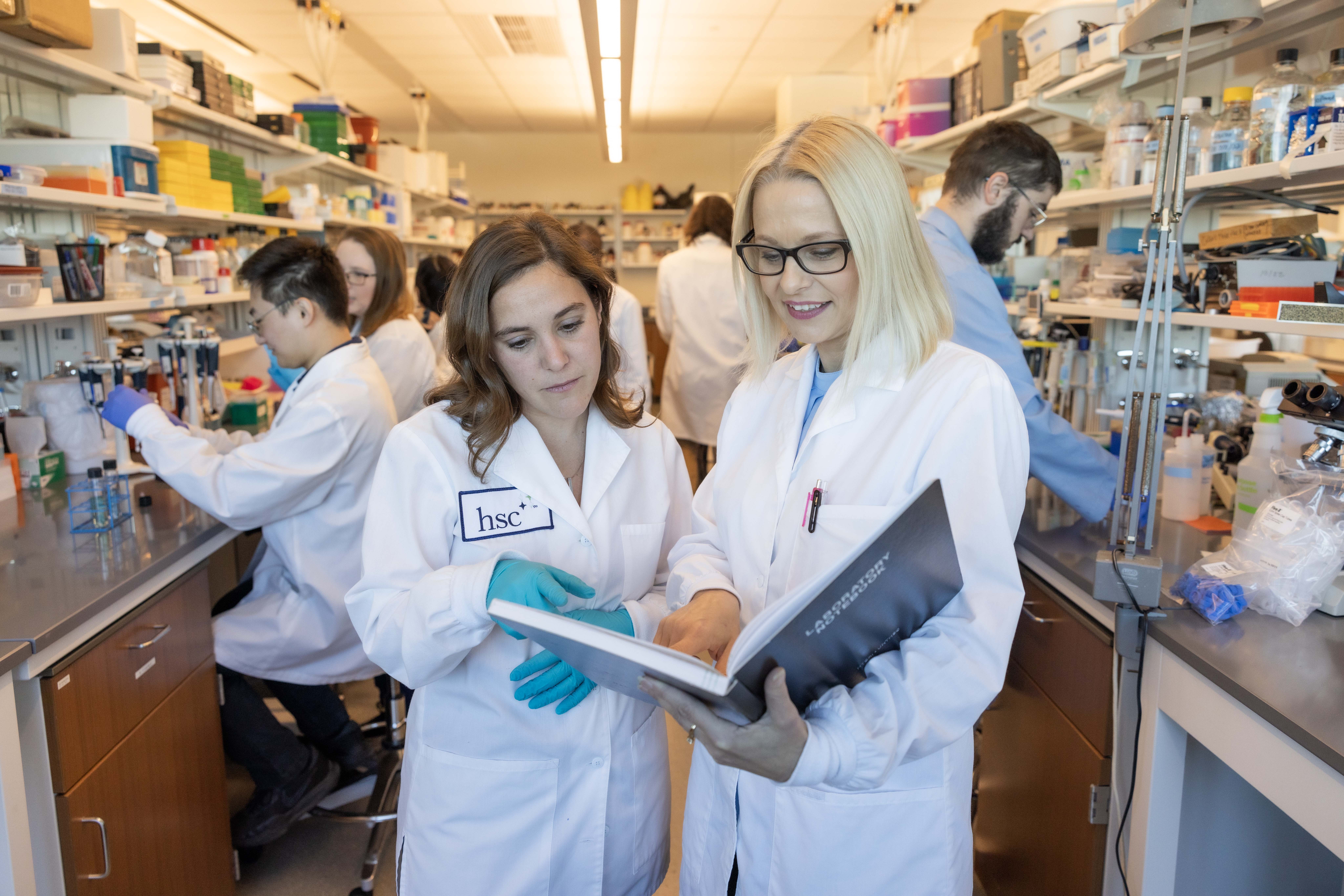 Photo of two female medical student researchers in white medical jackets looking at a bookbinder. Behind them are other student and faculty researchers handling test tubes and equipment sitting at opposing counters positioned on either side of them.