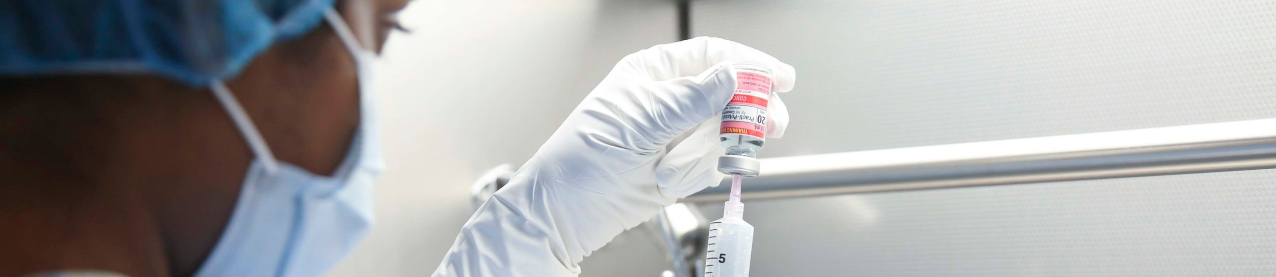 Top horizontal photo banner of a masked medical person wearing latex gloves using a syringe to pierce the upside-down pharmaceutical glass bottle being held