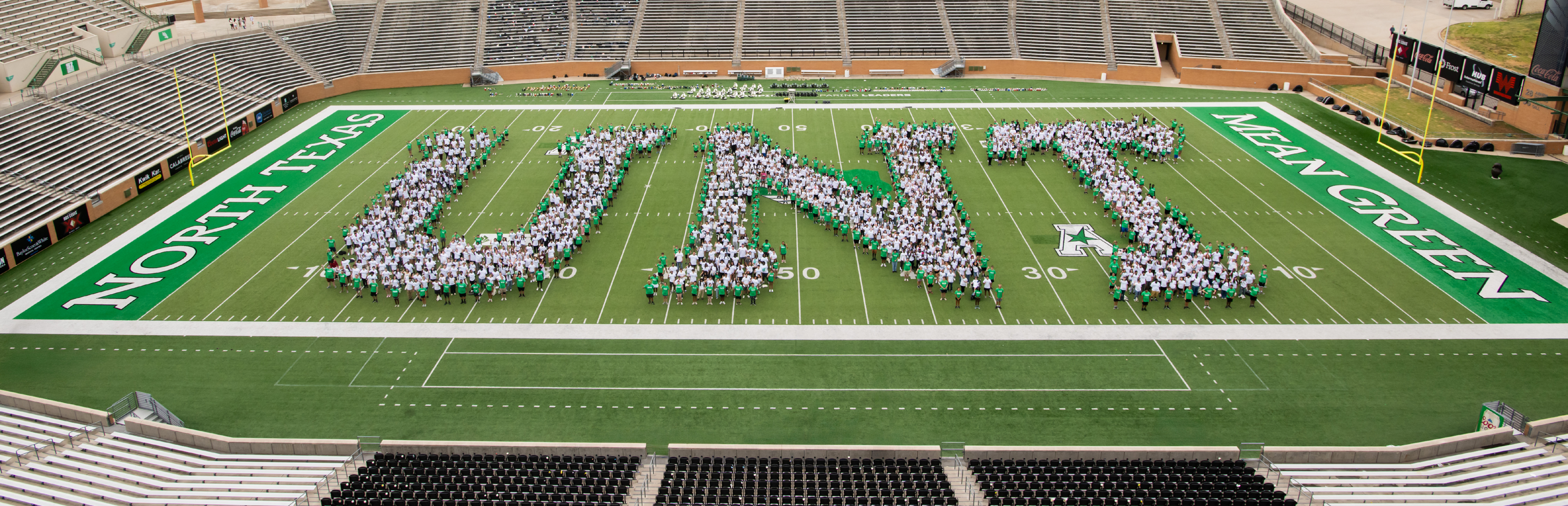UNT DATCU Mean Green Football Stadium with people in three groups on the field forming the separate letters of U N T in white clothes. There are a single row of people wearing green that form an outline edge to these letters.
