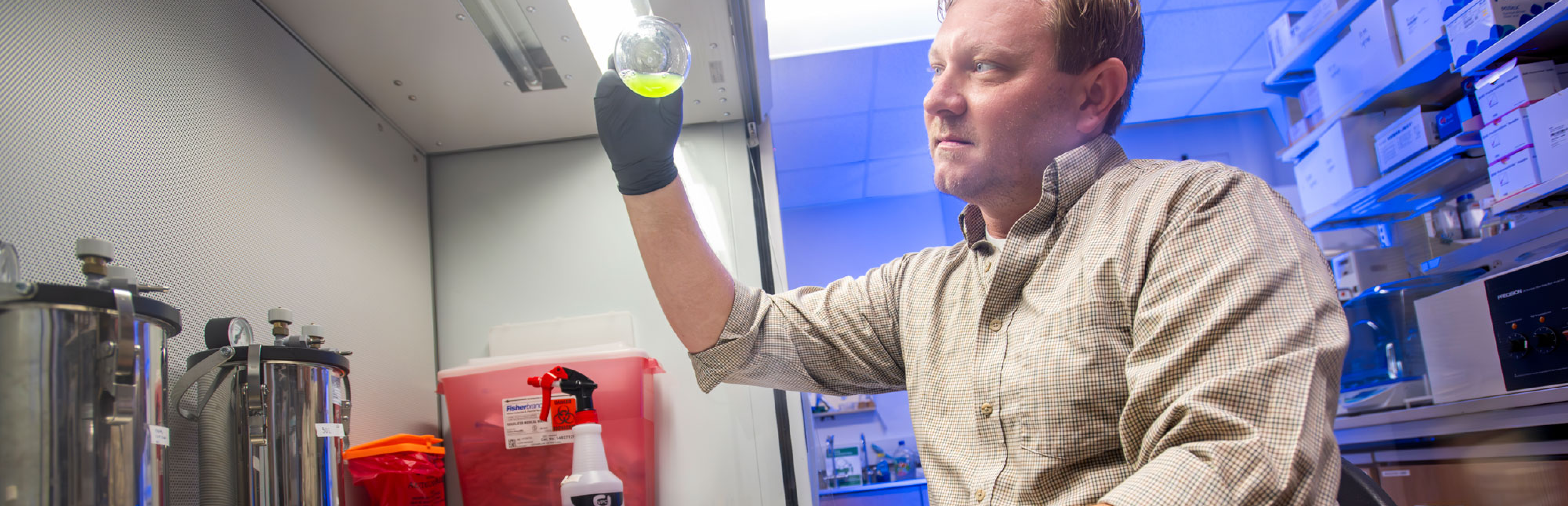 Photo of UNT's Calvin Henard looking at the bottom of a glass beaker with some lime green liquid that he is holding up to examine in his lab