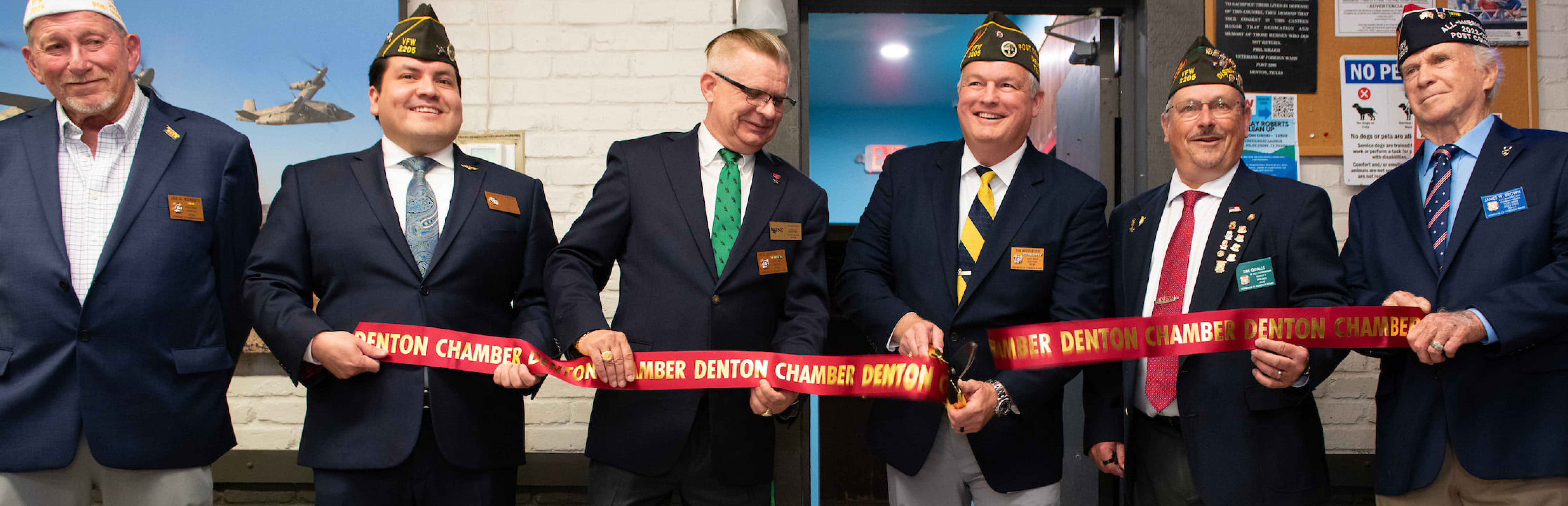 A photo showing a line of six veterans wearing suits, ties, and their vet hats are golding up a recent just cut Denton Chamber of Commerce red ribbon.