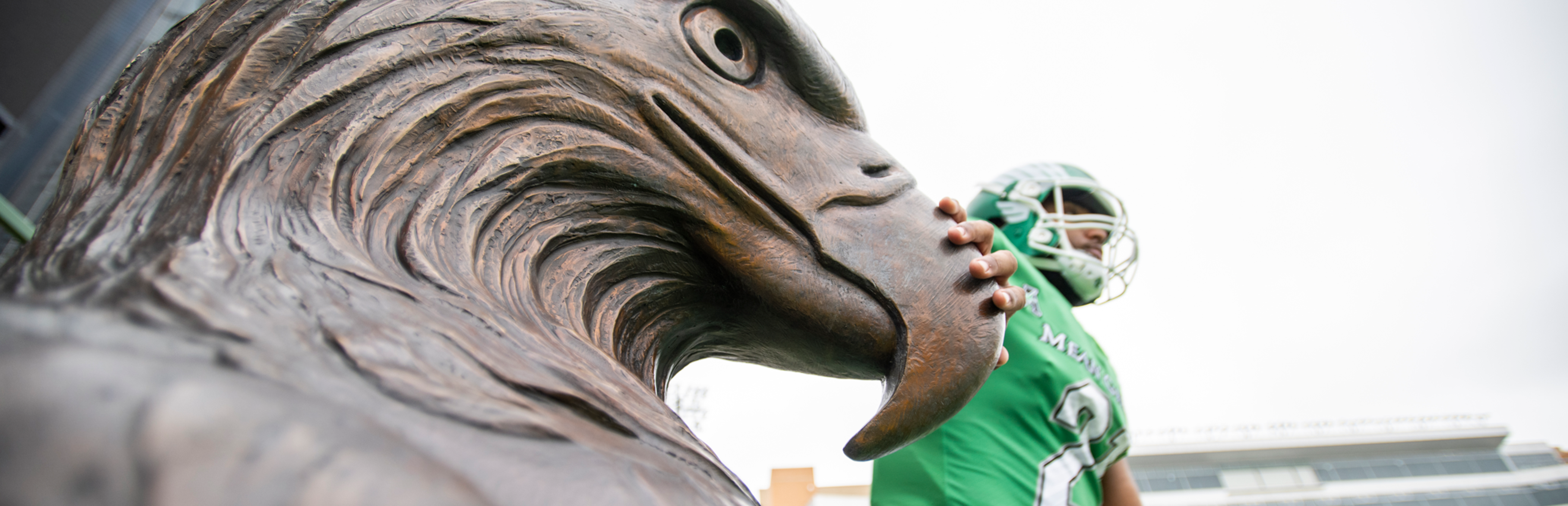 Photo of a close-up of the large head of a bronze eagle statue with a UNT football player in a green uniform behind it, with his hand rubbing its beak for luck as the player is about to enter the stadium field.