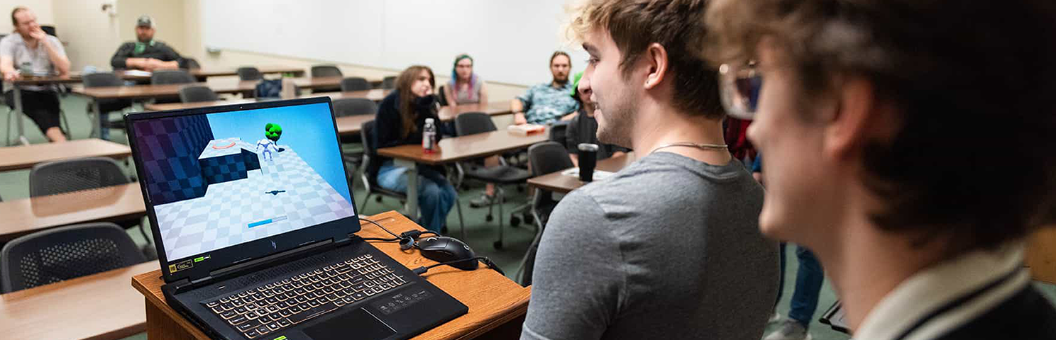 UNT students in a college classroom with two standing in the foreground by a lecture podium with an open laptop on it. The illuminated screen shows a digital 3D game graphic. The angle of the perspective is looking back into the room with some seated students.