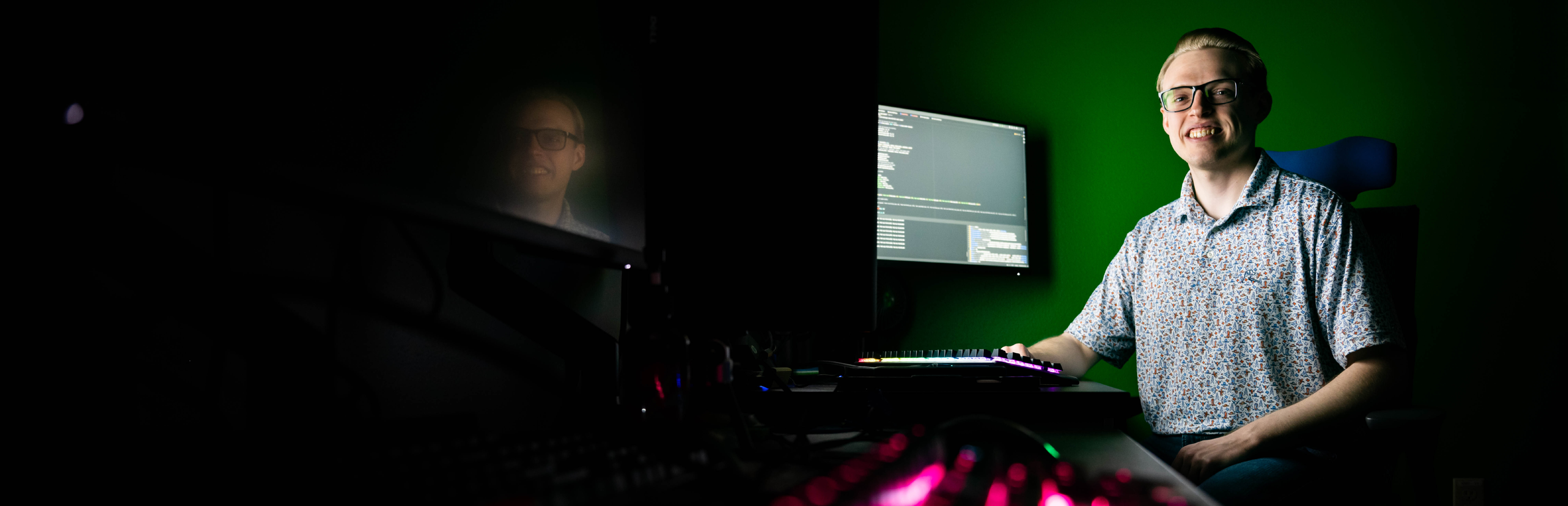 A horizontal photo of UNT's Garrett Cayce from the College of Engineering sitting in a dark room next to a computer console with lights and an illuminated computer screen in a darkened room
