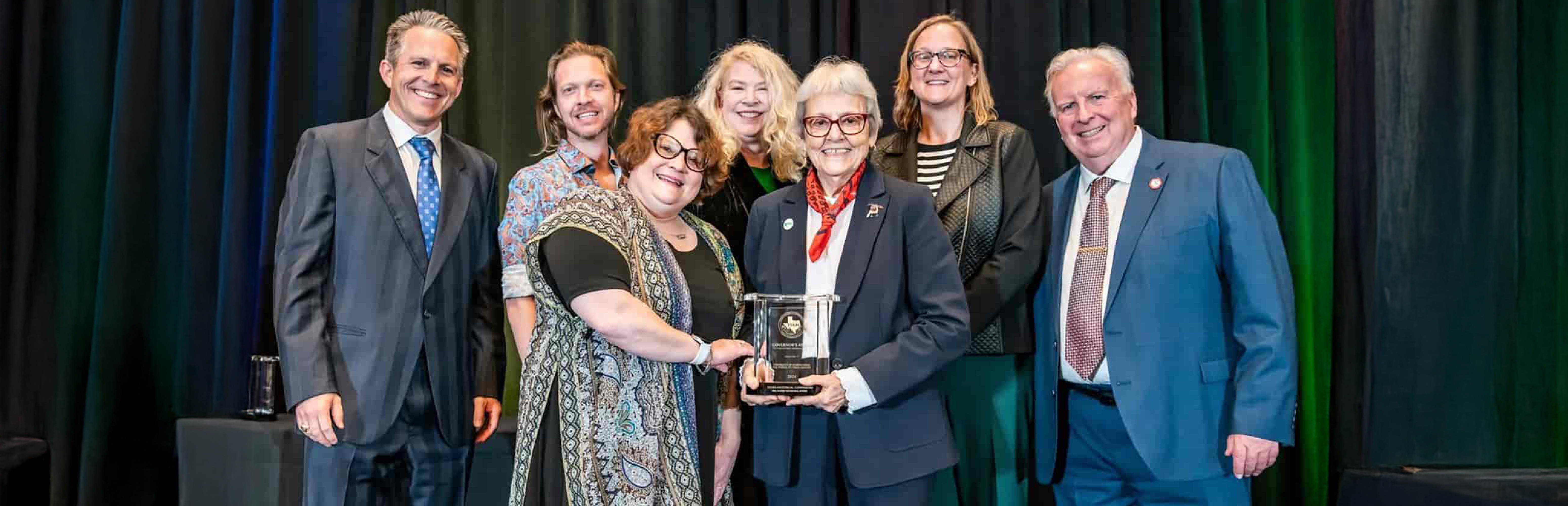The UNT Libraries staff stand on stage together for a photo op as one of them holds the top presented award at the 2025 Texas Historical Commission Governor's Award for Historic Preservation