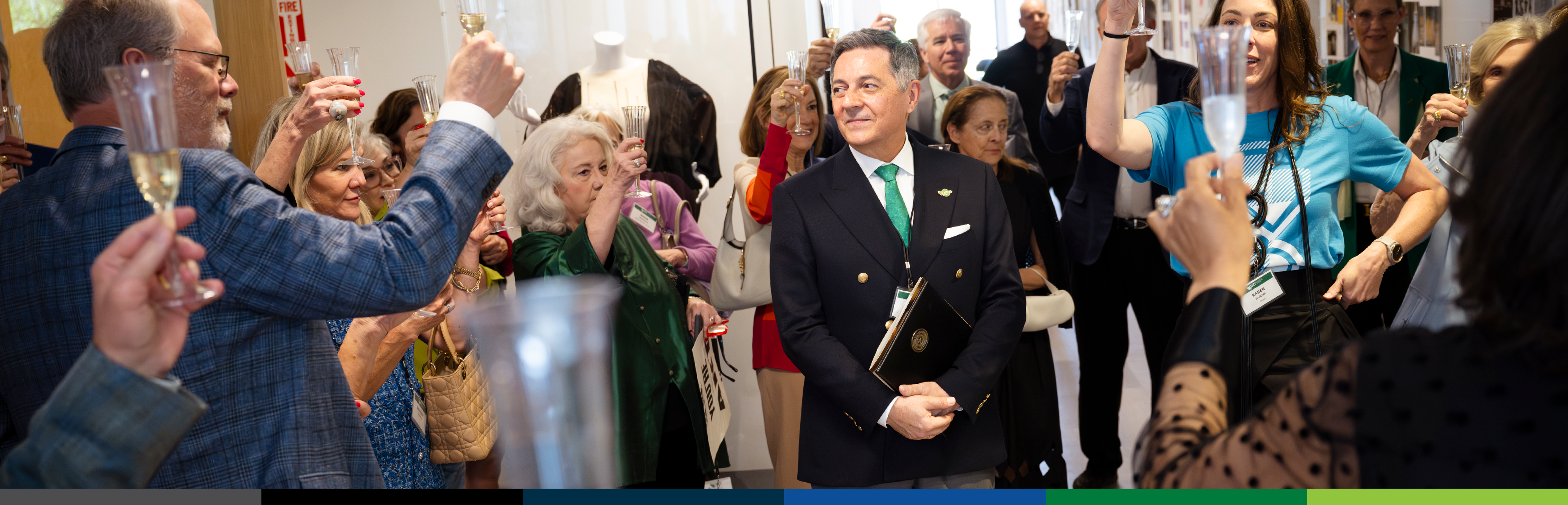 Michael W. Faircloth wearing a black-breasted sport coat, white shirt, and green tie standing in the middle of a crowd of UNT fashion supporters all raising their champagne glasses to toast him