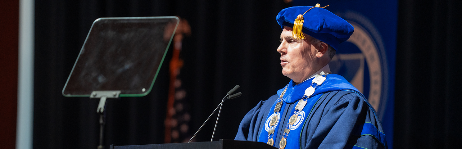 UNT Dallas President Dr. Warren von Eschenbach speaks at a podium in full academic regalia during his Investiture ceremony on May 1, 2026, announcing the Trailblazer Tuition Trust program.