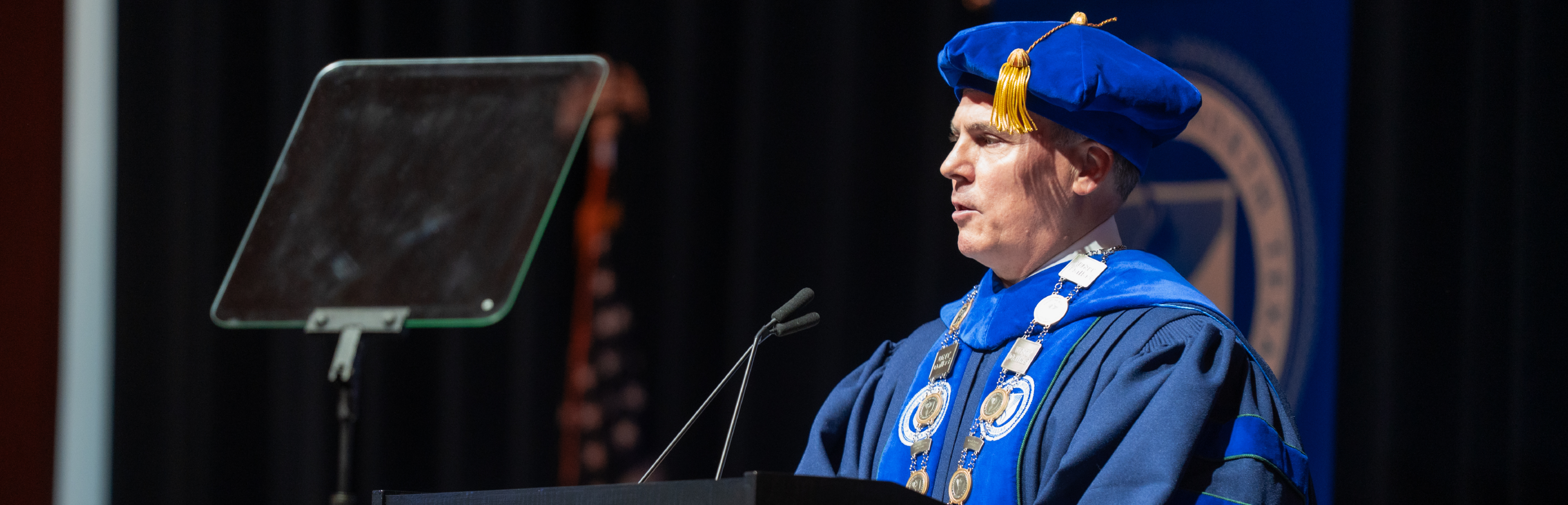 UNT Dallas President Dr. Warren von Eschenbach speaks at a podium in full academic regalia during his Investiture ceremony on May 1, 2026, announcing the Trailblazer Tuition Trust program.