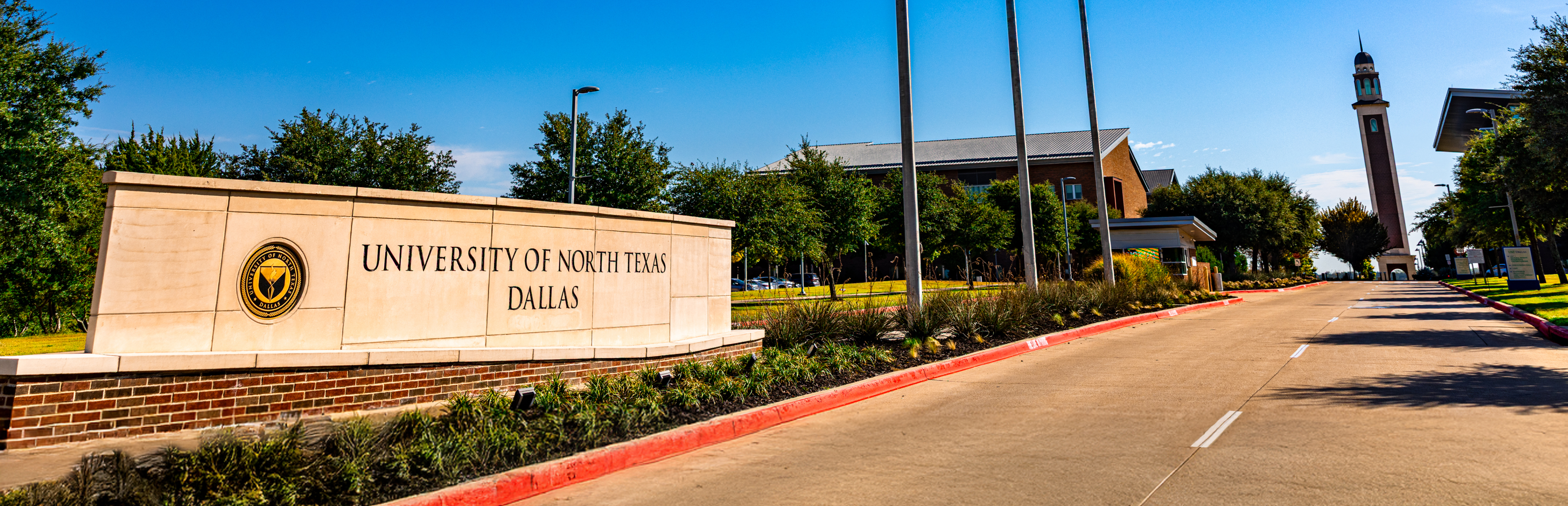 Photo of the UNT Dallas campus drive-up entry road. In the right foreground is a low entrance sign with the university's official seal and the engraved words "University of North Texas at Dallas" in black letters, along with campus buildings and the clock tower directly in the background.