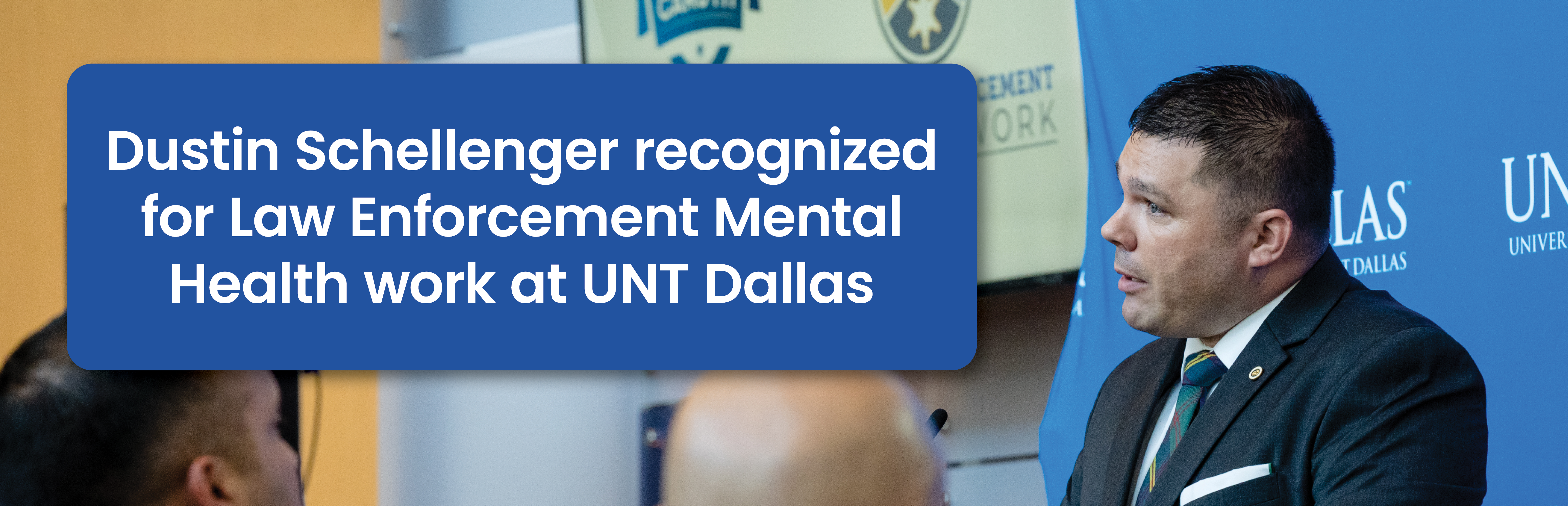The image features a man (Dustin Schellenger) speaking at an event. Included is a text banner that reads: "Dustin Schellenger recognized for Law Enforcement Mental Health work at UNT Dallas". Dustin Schellenger is wearing a dark suit with a white shirt and a tie. He is addressing an audience. There are a few people visible in the foreground, seen from behind.