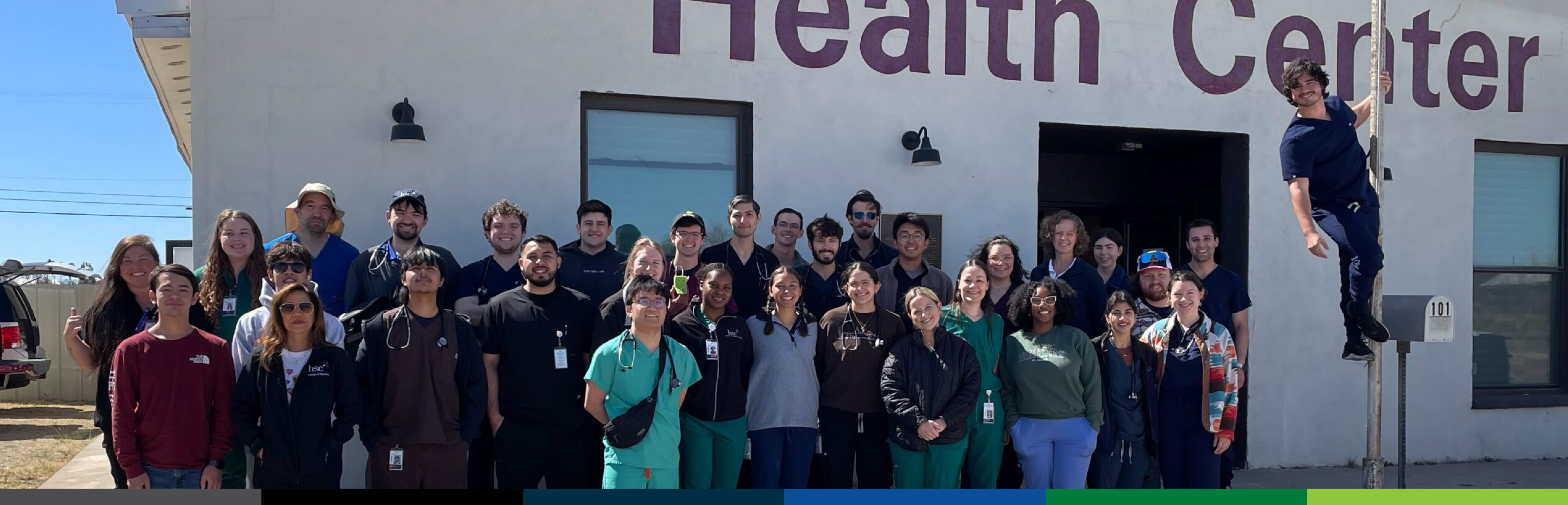 Photo of a group of 33 UNT Health TCOM students posing outside in front of a local small-town healthcare center, with one person playfully climbing a flagpole.