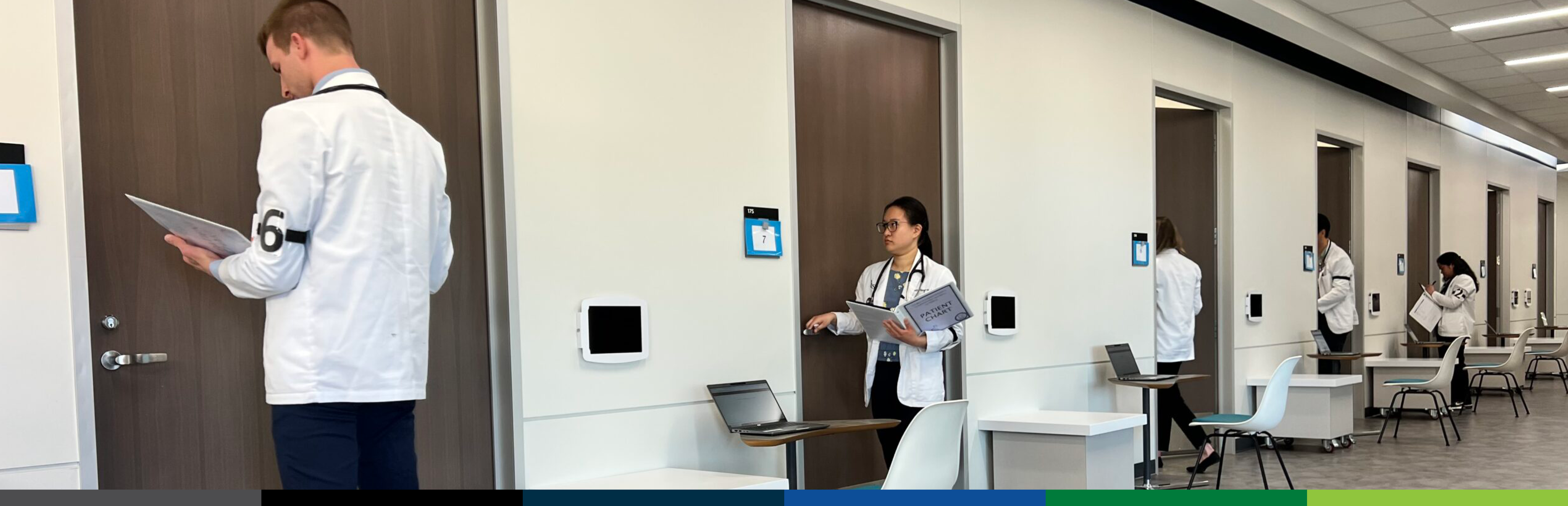 Photo of a group of 5 male and female UNT Health students in white coats prepare to enter numbered patient exam rooms in a clinical simulation center for assessment.