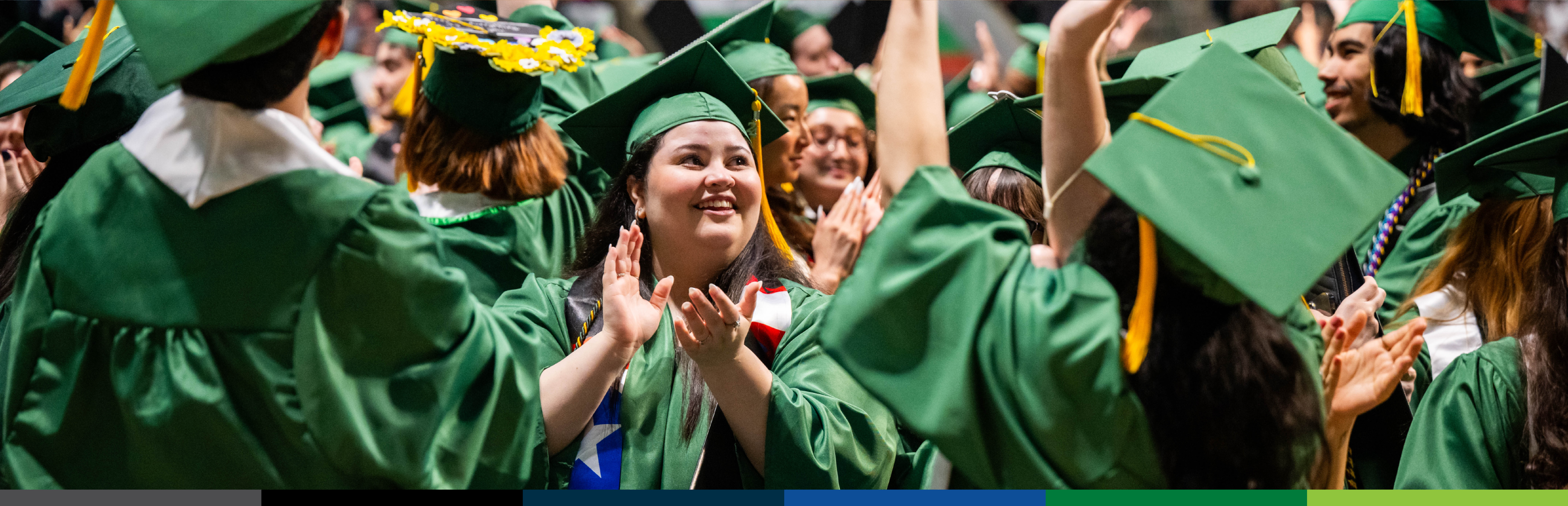 Photo of a group of graduates in green caps and gowns celebrate at a commencement ceremony. Almost everyone’s back is turned, facing the other way except for a female student in the center of the composition whose face is visible. She is looking off to the right side and happily applauding.